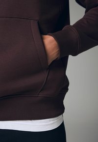 Sudadera marrón con un bolsillo tipo canguro, puños y dobladillo acanalados. Llevada sobre una camisa blanca, mostrando un diseño casual y texturizado.
