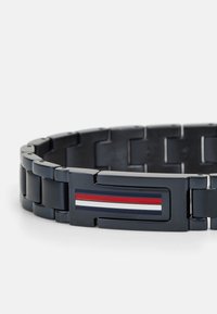 Mustmust bracelet, millede omavahel seotud lingid. Omab ristkülikukujulist lukku, millel on sinised, punased ja valged triibud. Sile tekstuur.