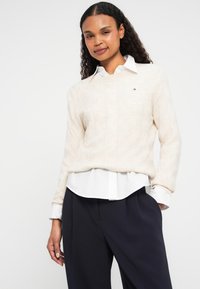 Beige gebreide trui met een gemarmerd patroon, gedragen over een witte blouse met kraag. Gecombineerd met hoge, donkere broeken.