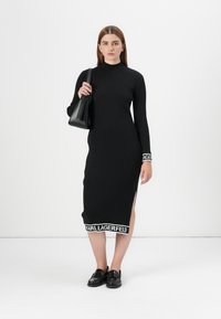 Svart ribbad stickad klänning med långa ärmar, sidoklipp och kontrasterande vit Karl Lagerfeld-logotyp i fållen. Modellen håller en svart väska.