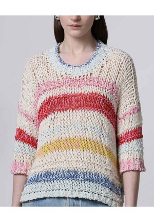 Vrouw draagt een losgebreide trui met brede horizontale strepen in blauw, geel, rood, roze en wit, gecombineerd met lichtblauwe spijkerbroek.