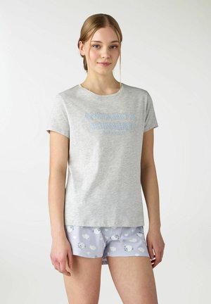 Giovane donna che indossa una maglietta grigio chiaro con la scritta "Andiamo a dormire" e pantaloncini azzurri con motivi di nuvole e pecore, in piedi all'interno.