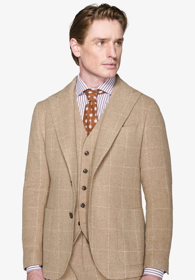 Beige villakangas puku, jossa on tarkistuskuvio, kiilakaarre, kuuden napin liivi ja ruskea pilkullinen solmio raidallisen paidan päällä.
