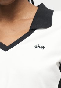 Κοντινό πλάνο ενός ατόμου που φοράει λευκό πουκάμισο με μαύρο γιακά και τη λέξη «obey» κεντημένη στο στήθος.