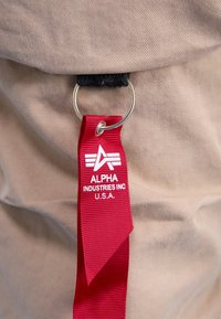 Czerwona metka z białym napisem i logo "Alpha Industries Inc U.S.A.", przymocowana do beżowej tkaniny metalowym kółkiem.