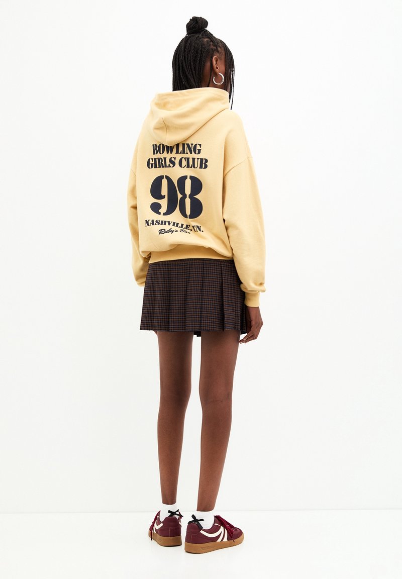 Jeune femme aux cheveux tressés porte un sweat à capuche jaune avec un texte noir et un numéro, une jupe à carreaux, des chaussettes blanches et des baskets bordeaux, debout, dos à la caméra.