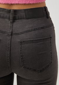 Black denim jeans feature a mid-rise design, subtle stitching details, and a single back pocket with a smooth texture. The waistband has a branded label.