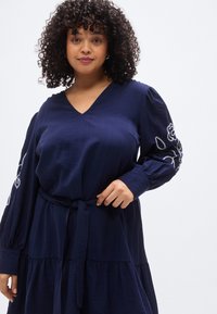 Φόρεμα σε ναυτικό μπλε με V-λαιμόκοψη, μακριά φουσκωτά μανίκια διακοσμημένα με λευκή κεντημένη ανθοδέσμη και δέσιμο στη μέση, κατασκευασμένο από ελαφρύ ύφασμα.