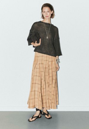 Černý pletený svetr s širokými rukávy, maxi sukně v béžovém károvaném vzoru a černé flip-flopy. Doplňky zahrnují dlouhý stříbrný náhrdelník a kovový náramek.