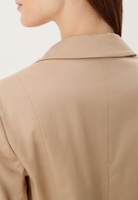 Close-up van de nek en schouder van een persoon die een beige blazer draagt met zichtbare stiksels en een klassieke kraag, tegen een witte achtergrond.