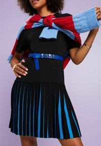 Femme portant une robe noire avec des rayures verticales bleues sur une jupe plissée, une ceinture bleue, et un pull rouge et bleuposé sur les épaules.