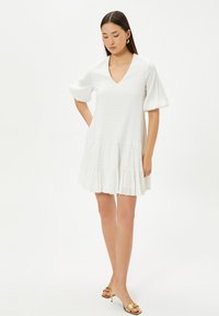 Robe blanche longueur genoux avec un motif texturé, décolleté en V, manches évasées et ourlet à volants superposés. Portée avec des talons ouverts dorés.