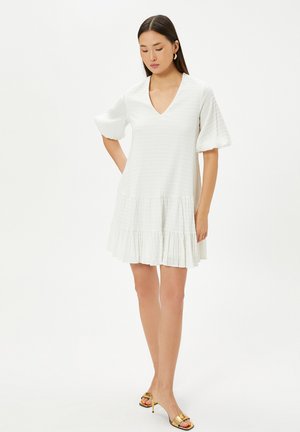 Robe blanche longueur genoux avec un motif texturé, décolleté en V, manches évasées et ourlet à volants superposés. Portée avec des talons ouverts dorés.