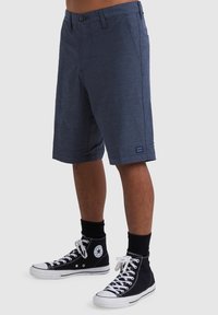 Uomo che indossa pantaloncini blu navy fino al ginocchio e scarpe Converse alte nere con calzini neri, in piedi su uno sfondo chiaro uniforme.
