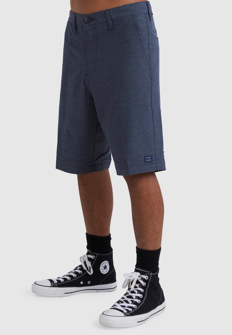 Navyblaue Shorts aus einem strukturierten Stoff, mit einem Knopfverschluss, Seitentaschen und einem Logo-Patch nahe dem Saum; kombiniert mit schwarzen High-Top-Schuhen.