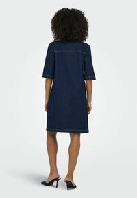 Denimklänning i mörkblått med rak form, korta ärmar, synlig sömnad och fåll precis ovanför knäet. Bärs med klackar.