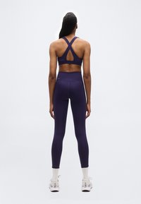 Γυναίκα που στέκεται γυρισμένη προς τα πίσω, φοράει αθλητικό σουτιέν με σταυρωτό σχέδιο στην πλάτη σε σκούρο μοβ, εφαρμόζουσες ψηλόμεσες κολάν στο ίδιο χρώμα, άσπρες κάλτσες και αθλητικά παπούτσια.