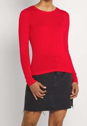 Osoba ubrana w dopasowany czerwony top z długim rękawem i czarną mini spódnicę z denimu z postrzępionym dołem na jednolitym tle.