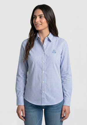 Mujer con camisa de cuadros azul y blanca abotonada con cuello y logo bordado en el pecho, combinada con vaqueros azules.