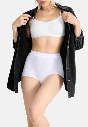 Kobieta ubrana w białe majtki z wysokim stanem i biały sportowy biustonosz, trzymająca rozpiętą czarną koszulę na jednolitym tle.