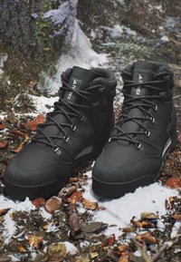 Stivali da trekking neri con lacci su terreno boschivo innevato con foglie, ramoscelli e tronco d'albero sullo sfondo.