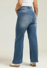 Siniset farkut, joissa on leveä lahdesign, taka-taskut ja koristelut sivuilla. Kangas on kuluneen näköinen.