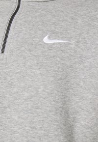 Grå zip-up sweatshirt i texturerad väv, med en vit Nike-logotyp på vänster sida av bröstet. Svart dragkedja.