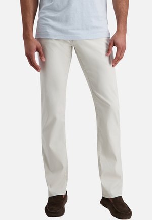 Mand iført lyse beige bukser med lige ben og brune ruskindssko, stående med hænder afslappet langs siden.