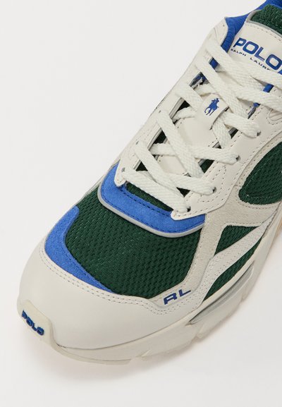 Grønn og hvit sneaker med mesh-overdel, blå detaljer og teksturerte materialer. Har snøring og en fremtredende logo på siden.