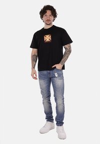 T-shirt nera a maniche corte con un grafico centrale rosso e oro, abbinata a jeans chiari strappati e sneakers bianche.