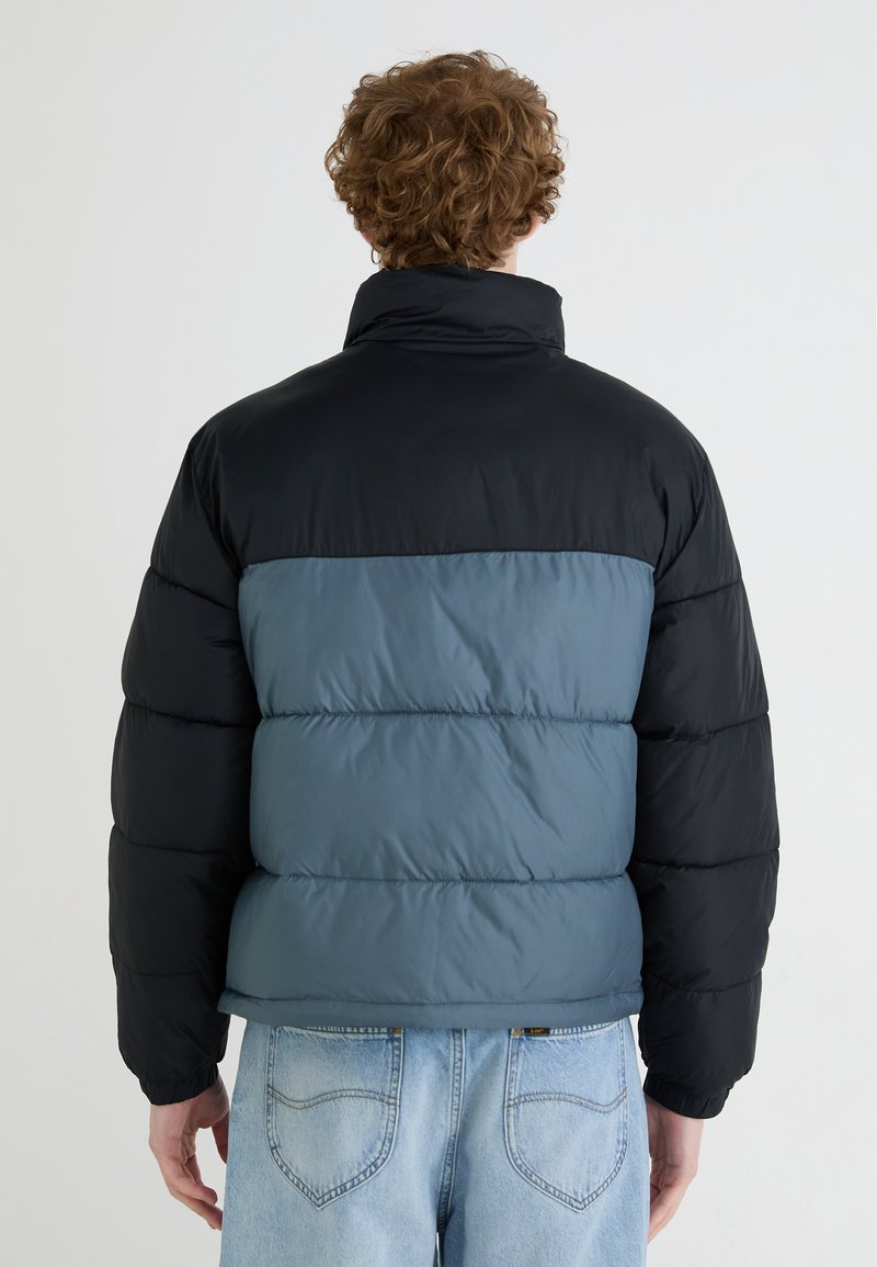 Doudoune en noir et bleu, avec un col montant, un design matelassé et des poignets élastiques. Le matériau semble lisse et légèrement brillant.