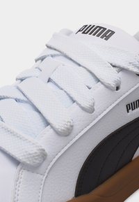 Λευκό αθλητικό παπούτσι Puma με χοντρές υφασμάτινες κορδόνια, μαύρο λογότυπο Puma στο πλάι και καφέ λάστιχο σόλα, κοντινή λήψη στην περιοχή των κορδονιών.