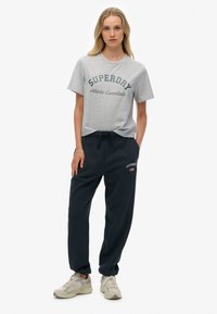 Camiseta de algodón gris claro con estampado "SUPERDAY Athletic Essentials". Pantalones de chándal azul marino oscuro con cintura elástica y detalle del logo en el bolsillo.