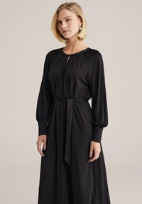 Fekete, hosszú ujjú ruha kulcslyuk nyakkivágással, derékban megkötős és lágy, áramló anyagból. Puffadt ujjakkal és lazább sziluettel rendelkezik.