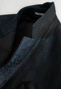 Tessuto a motivo nero e blu con una finitura testurizzata. Presenta un collo strutturato e una fodera liscia. Focus sul design del collo angolare.