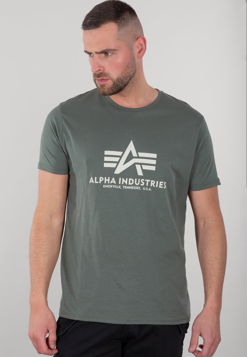 Olivově zelené tričko s krátkým rukávem vyrobené z měkké bavlny. Obsahuje bílé logo Alpha Industries se stylizovaným písmenem A a dvěma vodorovnými pruhy.
