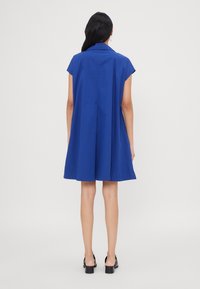 Donna di spalle che indossa un vestito blu cobalto, corto e a maniche corte, lungo fino al ginocchio, con sandali neri con tacco su uno sfondo neutro.