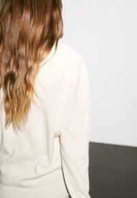 Maglione di cashmere crema, orlo e polsini a coste, vestibilità rilassata. Texture morbida, lunghi capelli castani mossi visibili, sfondo neutro con una superficie scura.