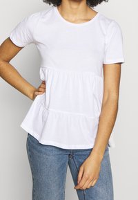 Bílý krátký rukáv tričko s kulatým výstřihem a peplum designem. Měkká látka, nařasený pas a volný střih. S ladícími modrými džíny.