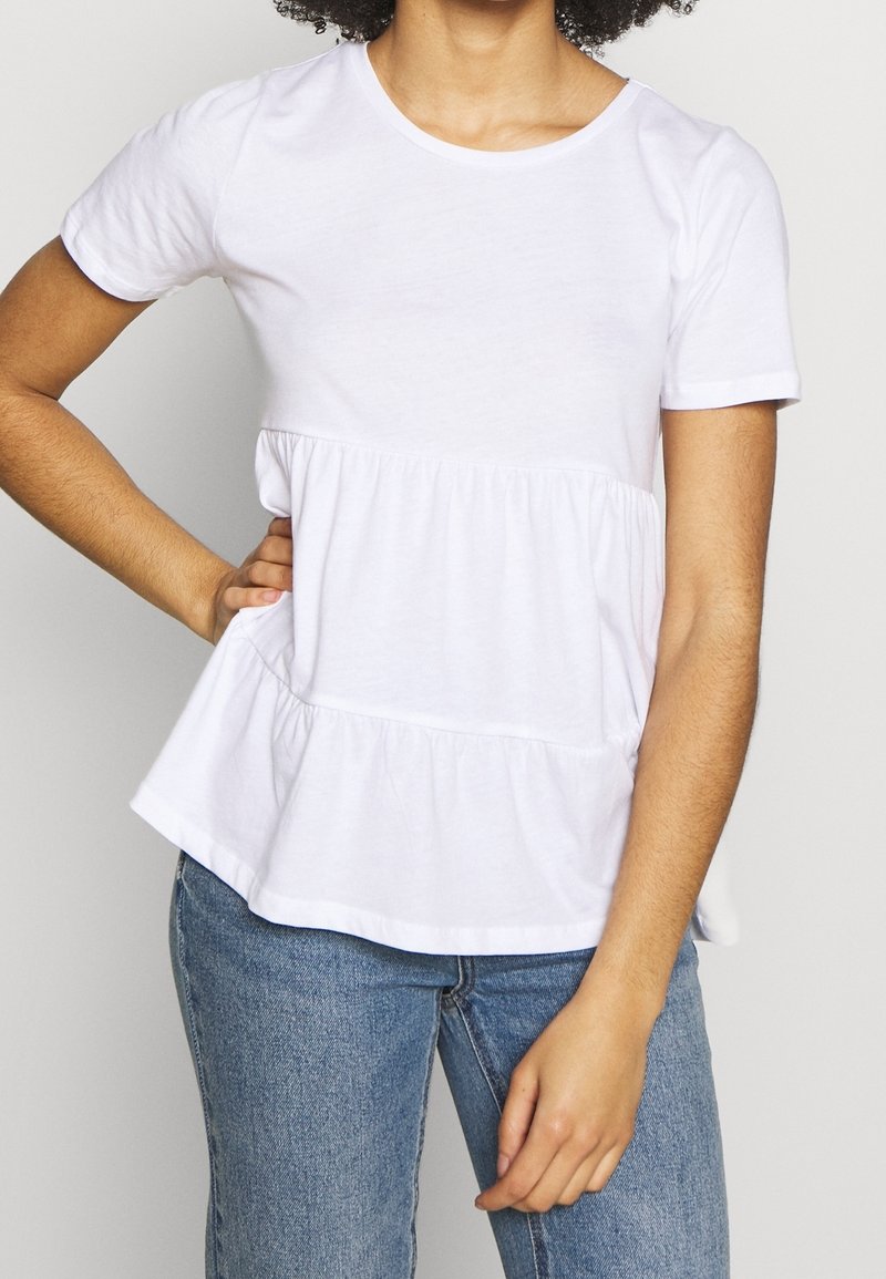 Bílý krátký rukáv tričko s kulatým výstřihem a peplum designem. Měkká látka, nařasený pas a volný střih. S ladícími modrými džíny.