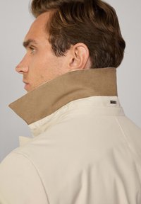 Chaqueta beige con un cuello alto en ante marrón. Presenta una textura sutil, un corte entallado y una pequeña etiqueta de la marca en el cuello.