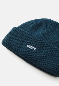 Tamsiai mėlynos spalvos megzta kepurė su riešinėmis tekstūromis. Su sulenktu kraštu ir baltu siuvinėtu "OBEY" logotipu.