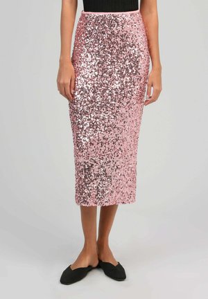 Femme portant une jupe midi en sequins rose brillant associée à des chaussures plates noires à bout pointu et un haut sans manches noir, debout devant un fond uni.