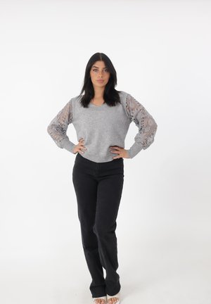Kobieta stojąca z dłońmi na biodrach, ubrana w szary sweter z koronkowymi rękawami i czarne spodnie na białym tle.