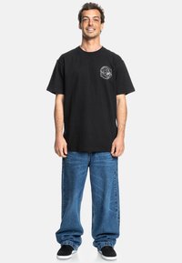 Black cotton t-shirt features a circular white graphic on the chest, paired with baggy blue denim jeans and black sneakers.