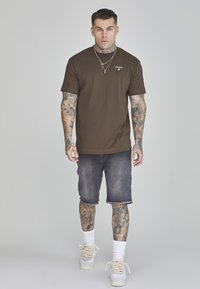 Camiseta de algodón marrón con cuello redondo, combinada con pantalones cortos de mezclilla oscura, calcetines blancos y zapatillas de deporte beige. Tatuajes notables en los brazos.
