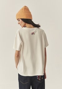 Kremowa bawełniana koszulka z krótkim rękawem, z kolorowym haftowanym napisem na górnej części pleców. Do tego ciemnoniebieskie jeansy z kwiatowym haftem.