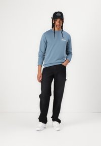 Lys blå hettegenser, svarte jeans, hvite sneakers og en svart caps med tekst, står foran en ensfarget bakgrunn. Lange fletter er synlige.