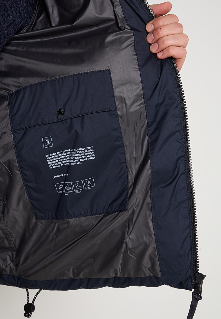 Das Innere der Marinejacke zeigt ein glattes, glänzendes Futter, einen Reißverschluss und eine Tasche mit Pflegeanweisungen und Angaben zur Stoffbeschaffung.