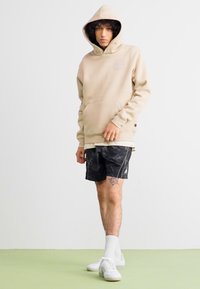 Felpa beige con cappuccio e tasca frontale, indossata su pantaloni corti neri con motivo. Il modello si trova su un pavimento verde, indossando calzini e scarpe bianche.